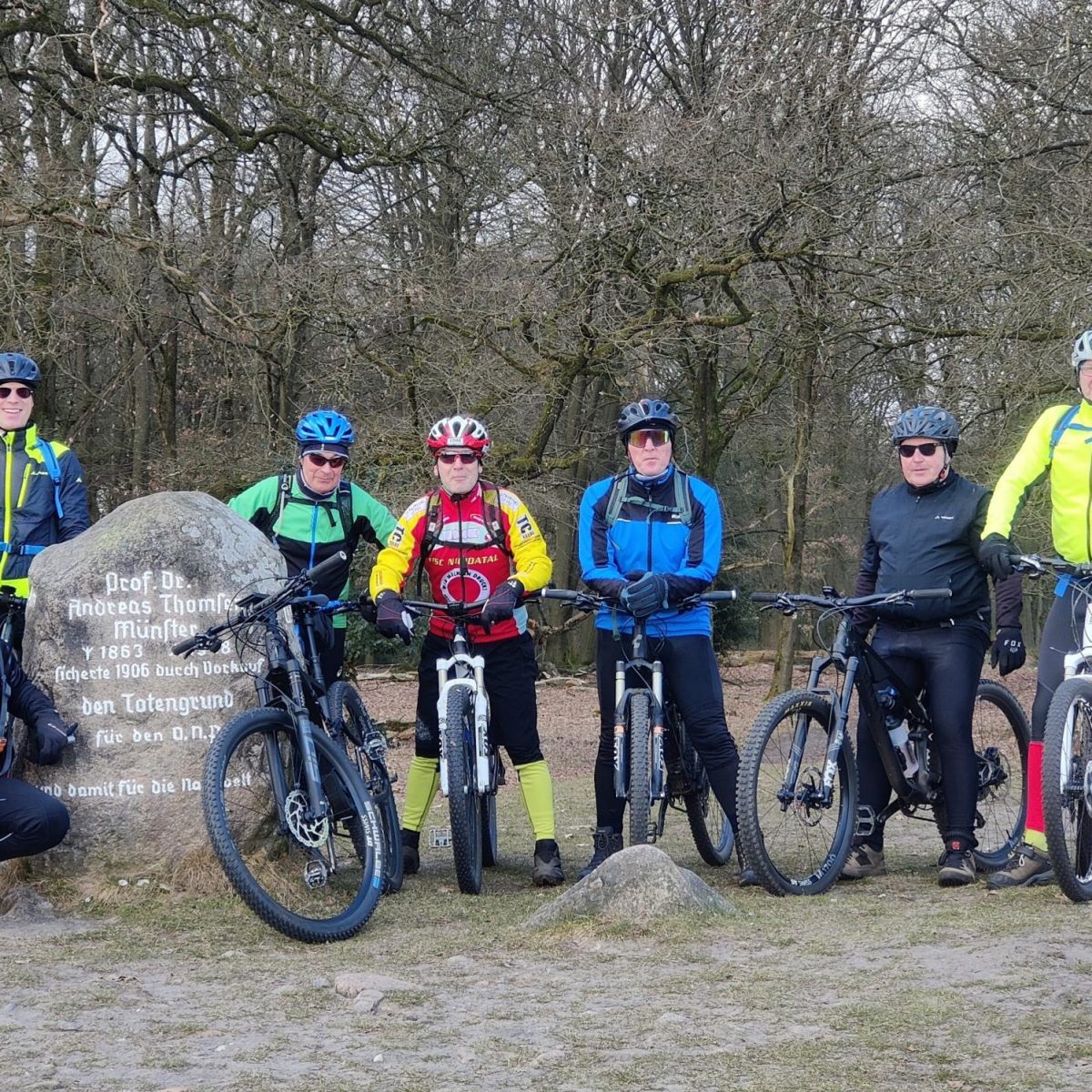 Eine Gruppe von 7 Männern in Mountainbike-Montur, die auf Ihren Fahrrädern sitzen und neben einem Felsen posieren.