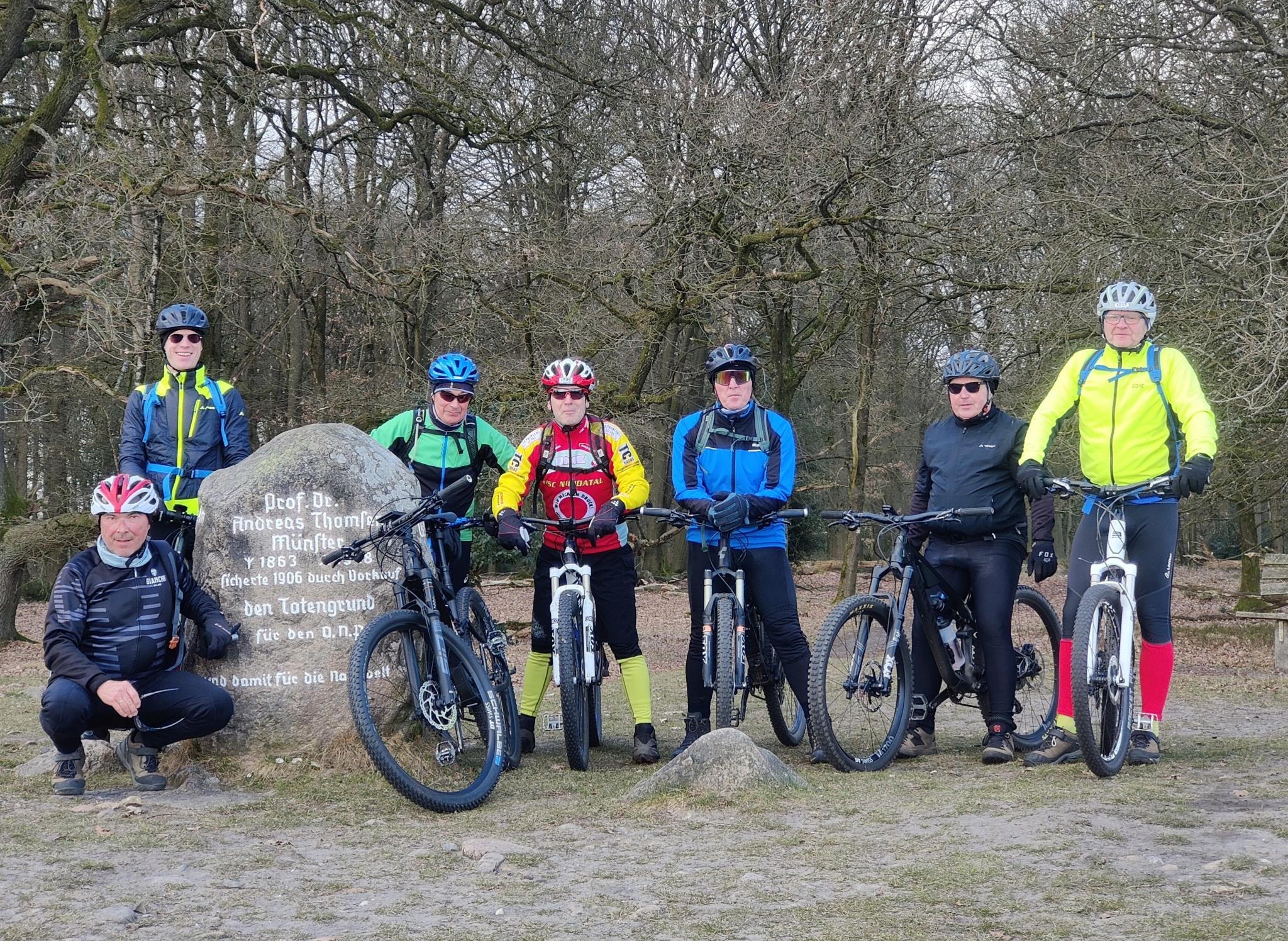 Eine Gruppe von 7 Männern in Mountainbike-Montur, die auf Ihren Fahrrädern sitzen und neben einem Felsen posieren.